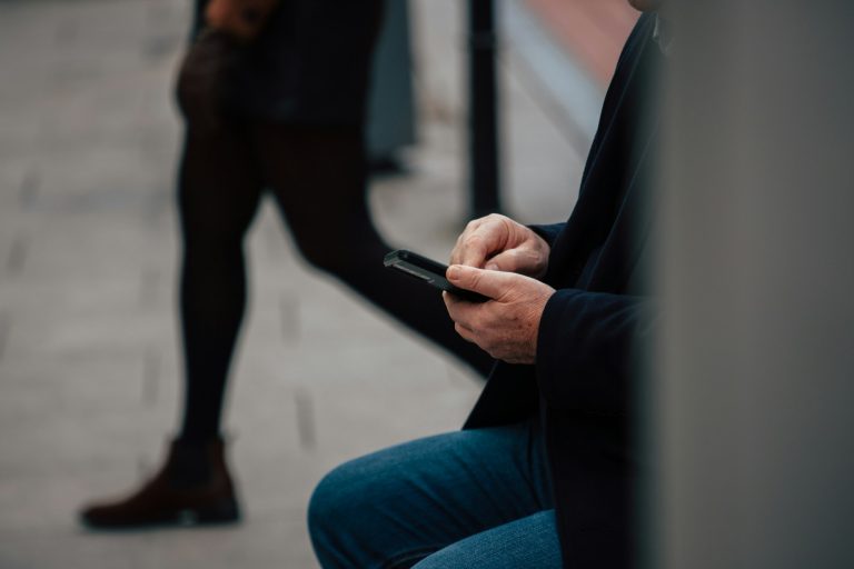 Man seated using a smartphone while a person walks past behind him on a city street