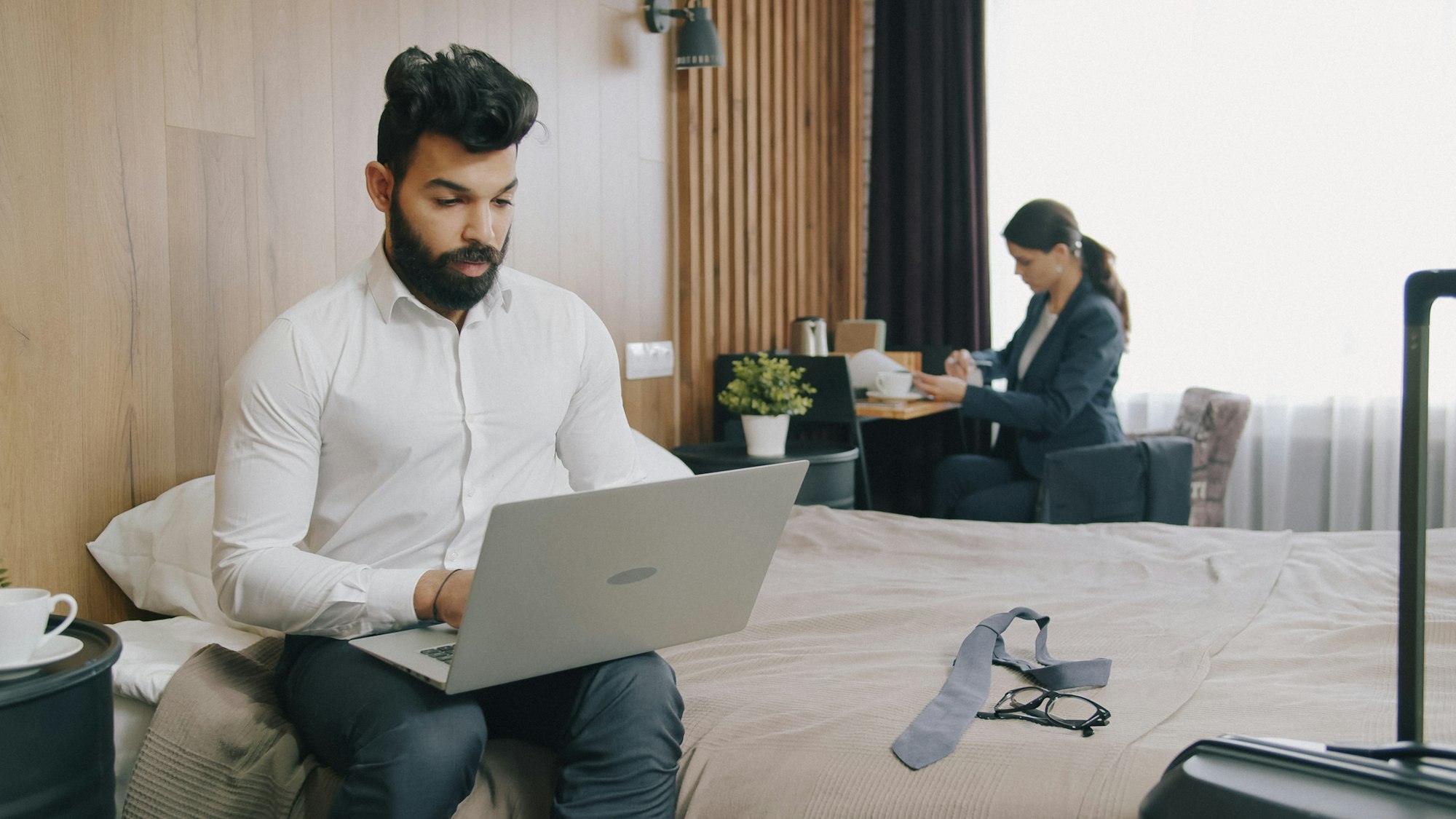 Man sitting on a hotel bed working on a laptop while a woman sits at a desk in the background