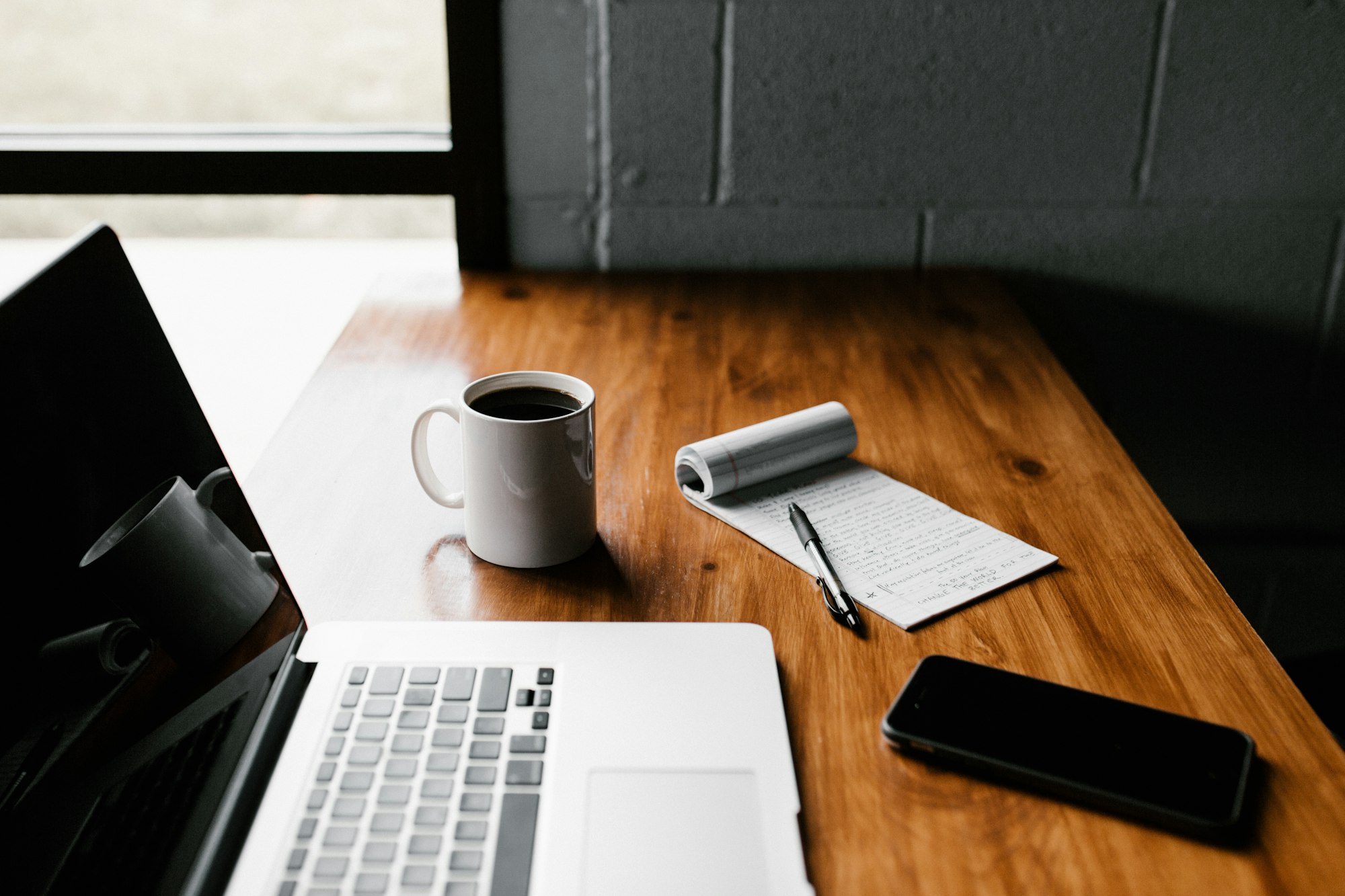 Laptop, a white mug of coffee, a notepad and a black smartphone on a wooden desk by a window