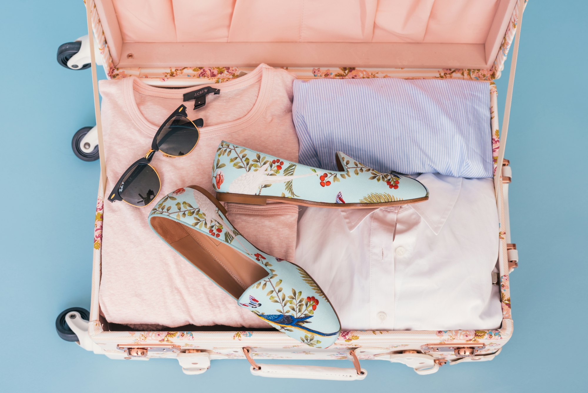 Open pink suitcase seen from above, packed with folded shirts, a pair of floral flats and sunglasses
