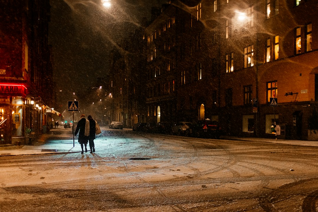 Two people walking along a snow-covered Stockholm street at night under glowing street lamps