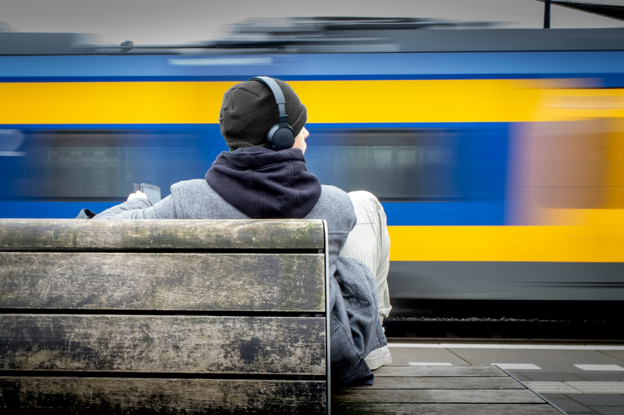 Person in a beanie and over-ear headphones sitting on a platform bench as a blue and yellow train blurs past